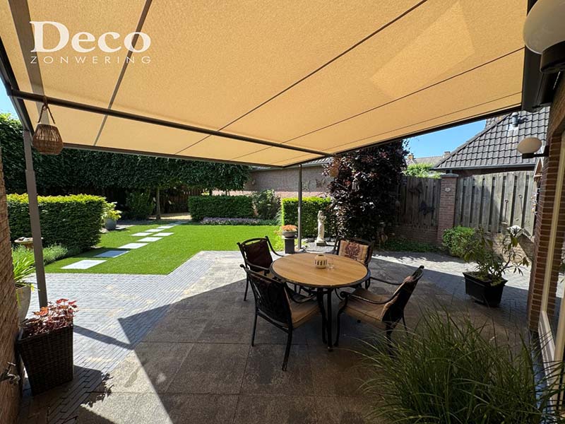 weinor-plaza-viva-overkapping-venhorst-uitzicht-tuin Binnenaanzicht van een Weinor Plaza Viva overkapping met crème-taupe doek, met zicht op een verzorgde tuin met gazon, stapstenen en hagen.