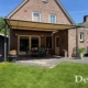 Totaaloverzicht van een Weinor Plaza Viva doek overkapping in grijsbruin met crème doek, geplaatst tegen de gevel van een vrijstaande bakstenen woning, met terras, gazon en beplanting.