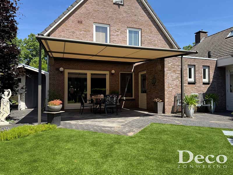 Totaaloverzicht van een Weinor Plaza Viva doek overkapping in grijsbruin met crème doek, geplaatst tegen de gevel van een vrijstaande bakstenen woning, met terras, gazon en beplanting.