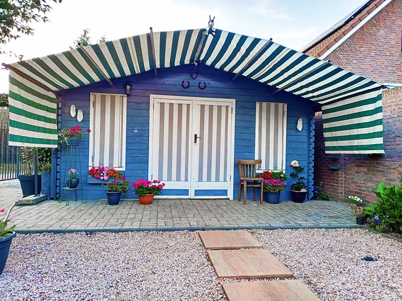 Hergebruikt groen-wit gestreept zonneschermdoek als duurzame zonwering bij een blauw tuinhuis Tuinhuis met hergebruikt gestreept zonneschermdoek in groen en wit, gespannen over het dak en voor de deuren en ramen als duurzame zonwering.
