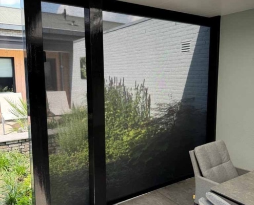 Windvaste Heroal solar screen zijkant overkapping Neergelaten windvaste Heroal solar screen aan de open zijkant van een overkapping in Heesch, met zicht op de tuin